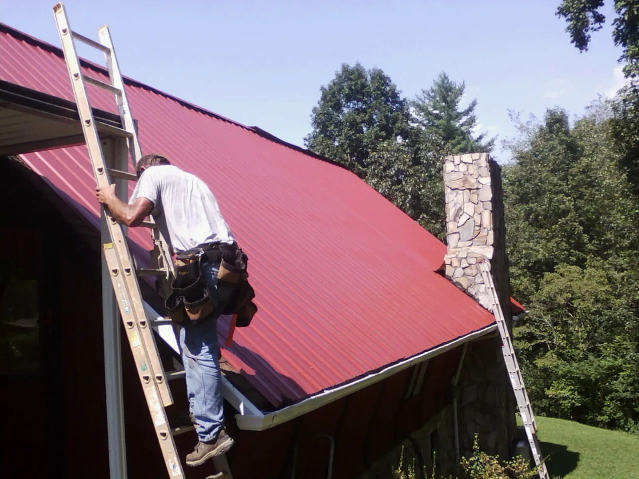 Roofer on a ladder beside a red metal roof and stone chimney — Soffit & Fascia Repair by Roofing Brothers in Leonardville